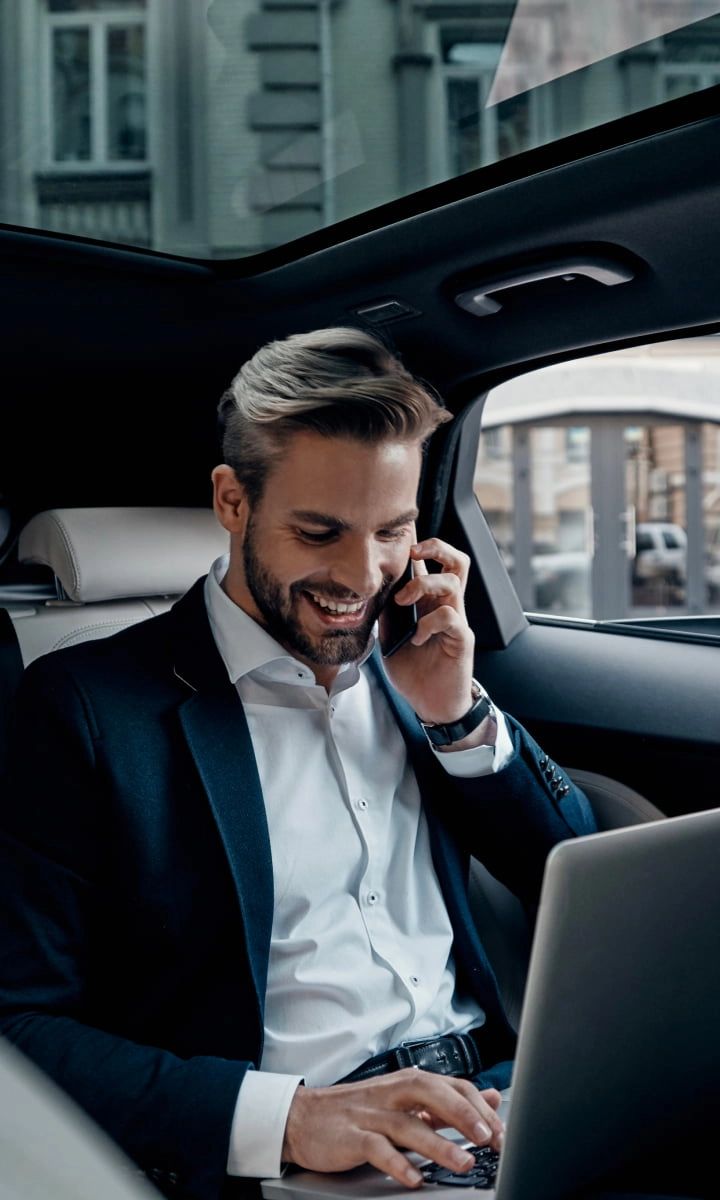 Executivo em fato a trabalhar no telemóvel e no computador no interior de um carro de luxo