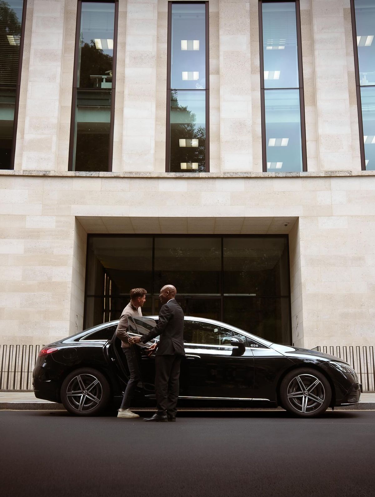 Motorista a receber um executivo junto a um carro preto à entrada de um edifício corporativo